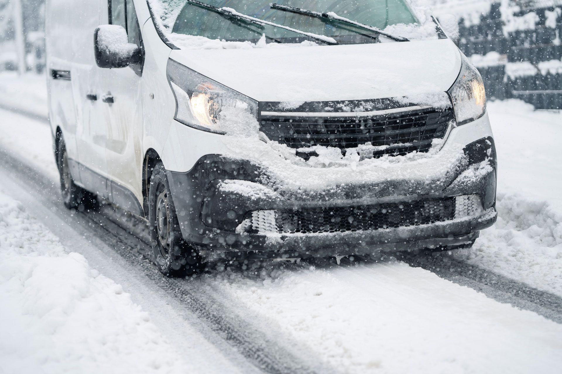 White van driving on a snow-covered road, headlights on, snow falling.