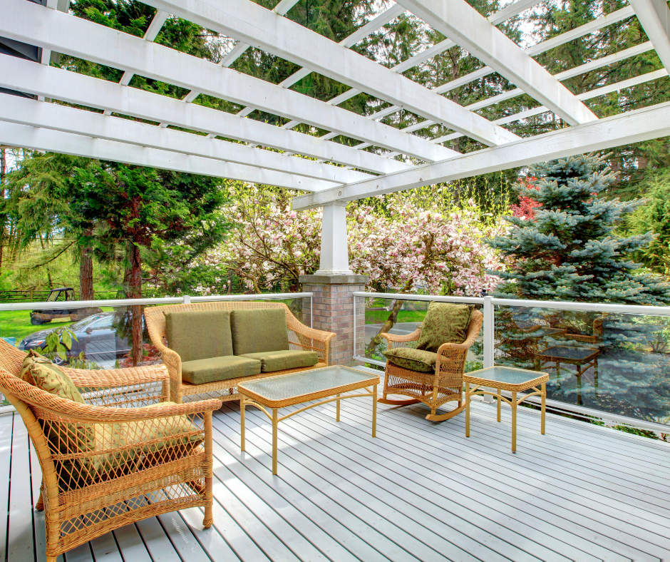 White-painted wooden deck with wicker furniture under a pergola. Lush green trees and flowers in the background.