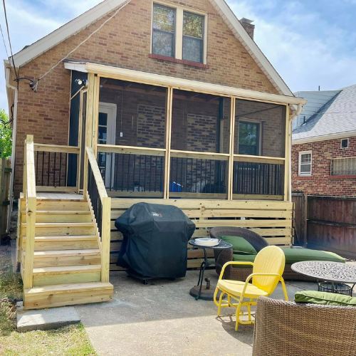 Backyard with a wooden deck, stairs, and screened porch attached to a brick house; patio furniture sits on the concrete patio.