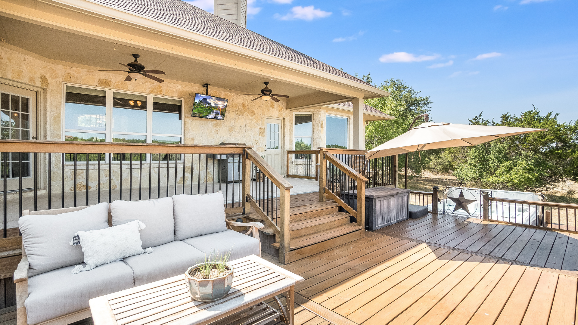 A wooden deck with seating, a hot tub, and an umbrella is attached to a stone house on a sunny day.