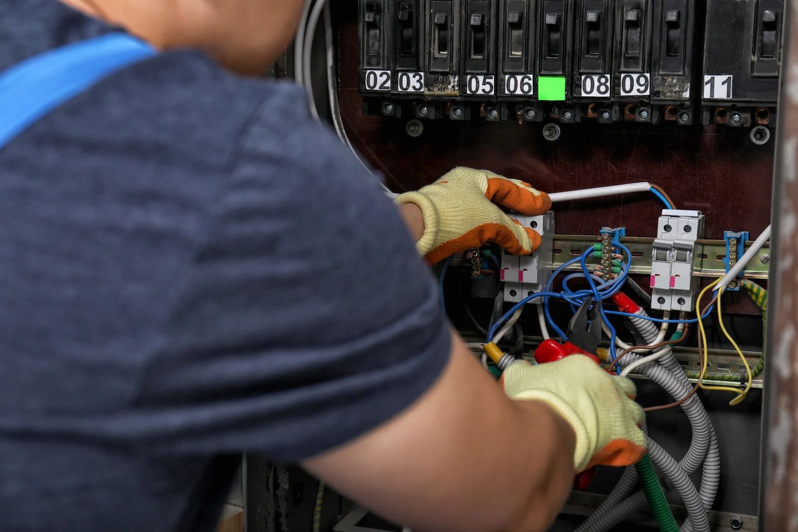 Electrician working on a circuit breaker panel, wearing gloves, using tools.