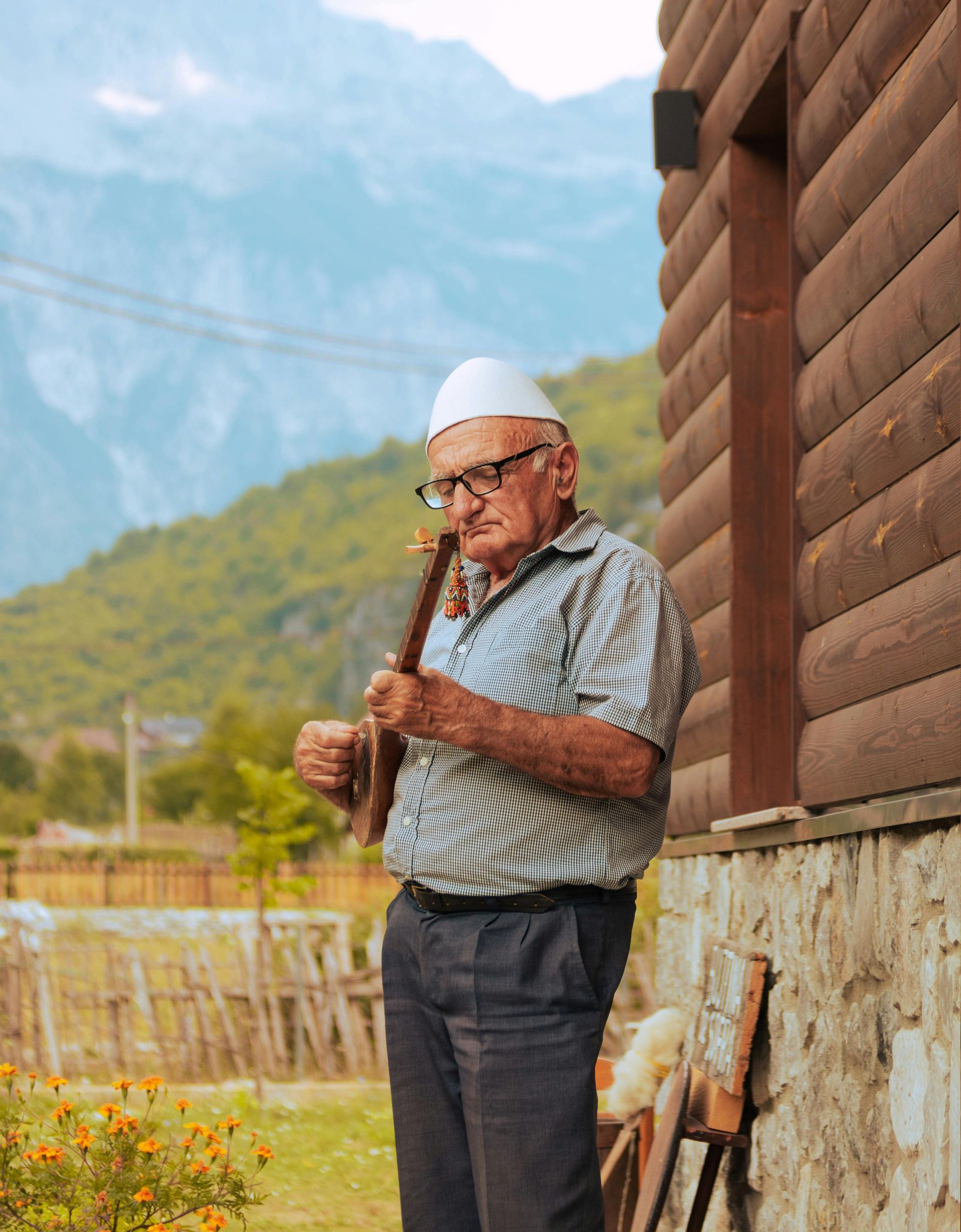 Un uomo anziano è in piedi davanti a una baita di tronchi e tiene in mano uno strumento tradizionale albanese