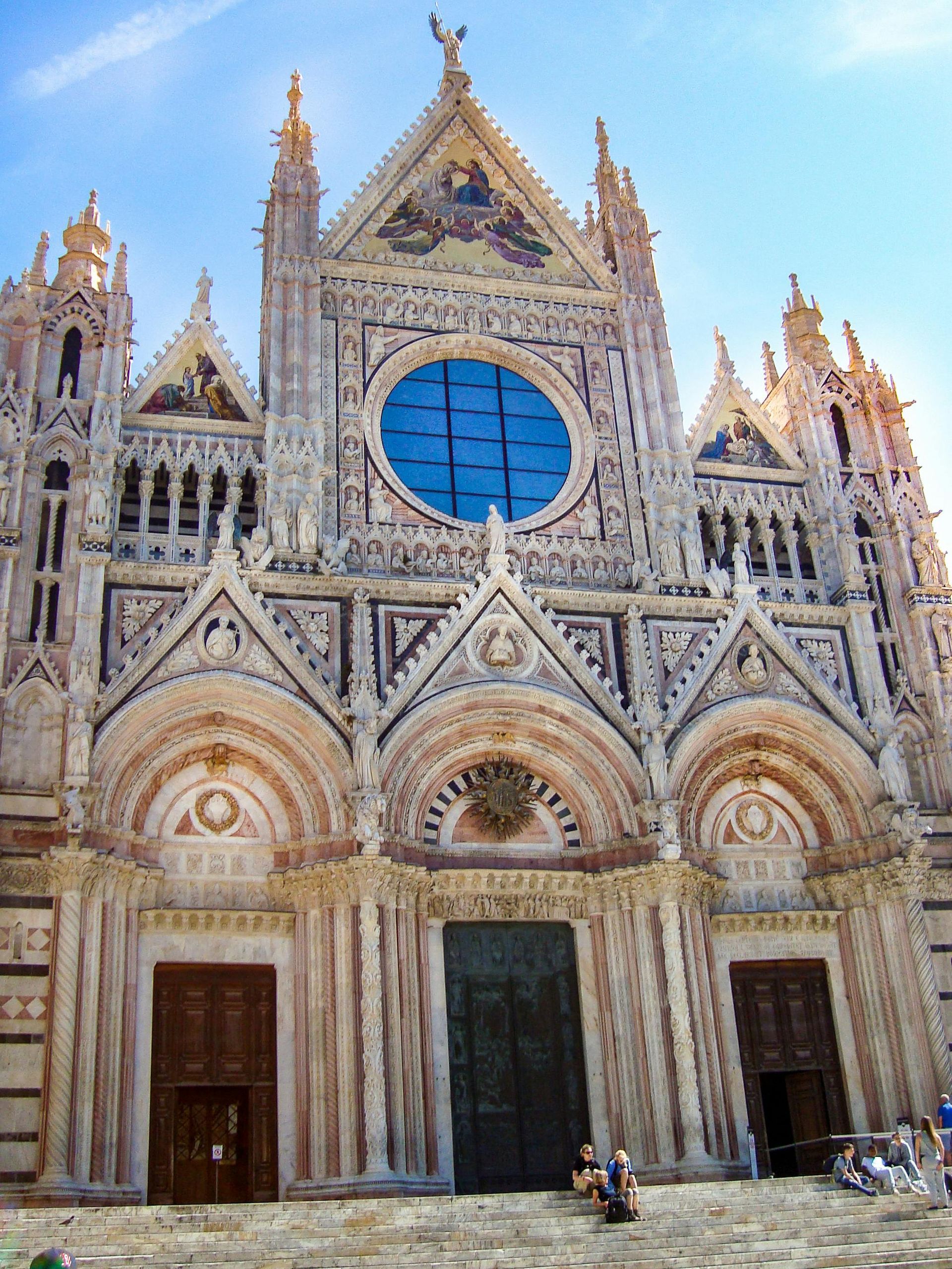 Facciata del Duomo di Siena con marmo bianco e verde, rosone e tre portali ad arco. Cielo azzurro.