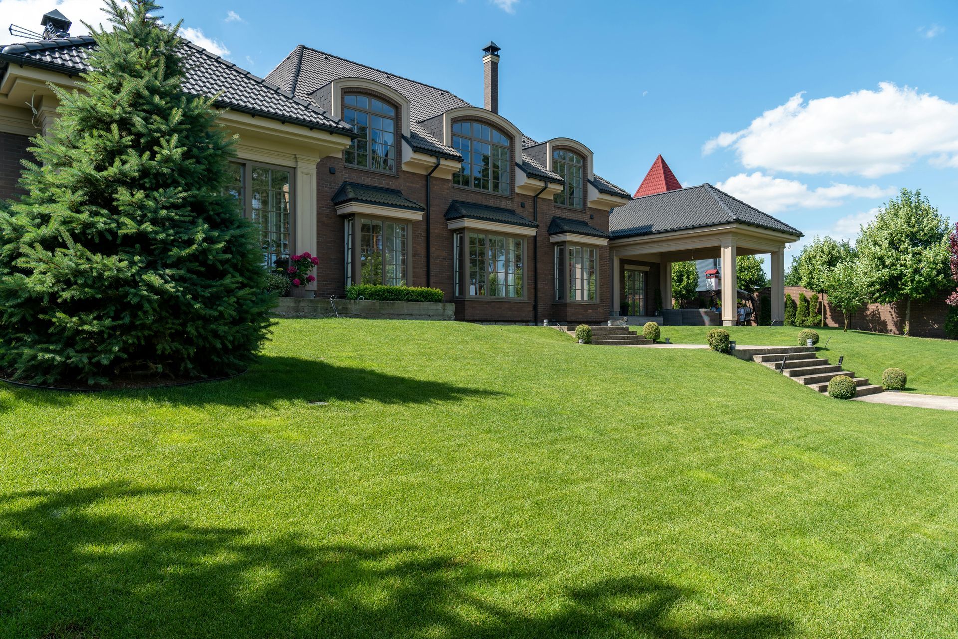 Large brick house with a green lawn, dark roof, and covered patio under a blue sky