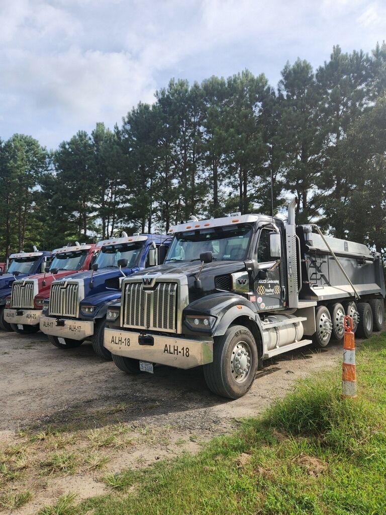 Blue dumpster on a truck bed, filled with debris, tilted in front of a house on a sunny day.