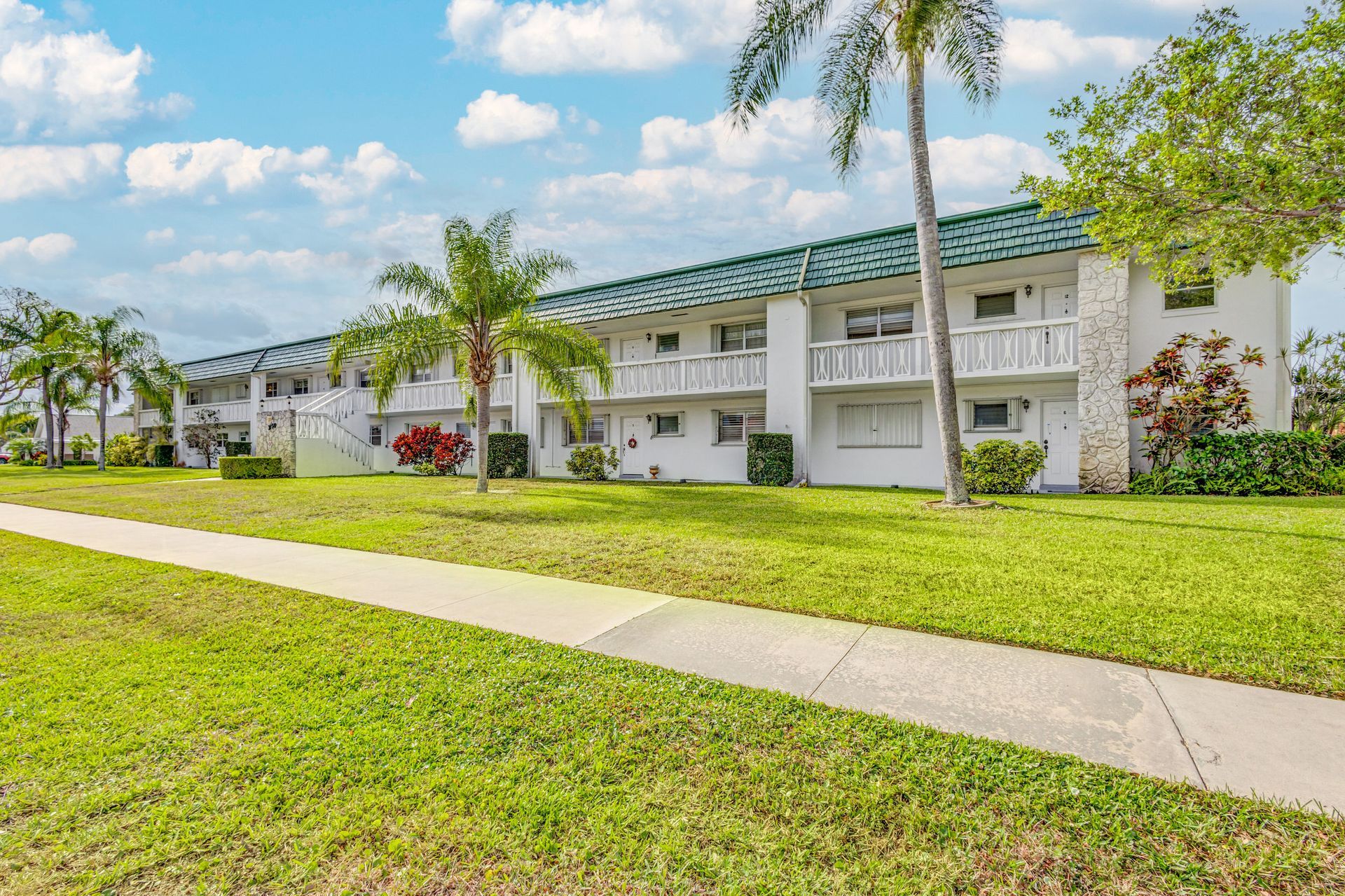 A white apartment building with a green roof is surrounded by lush green grass and palm trees.