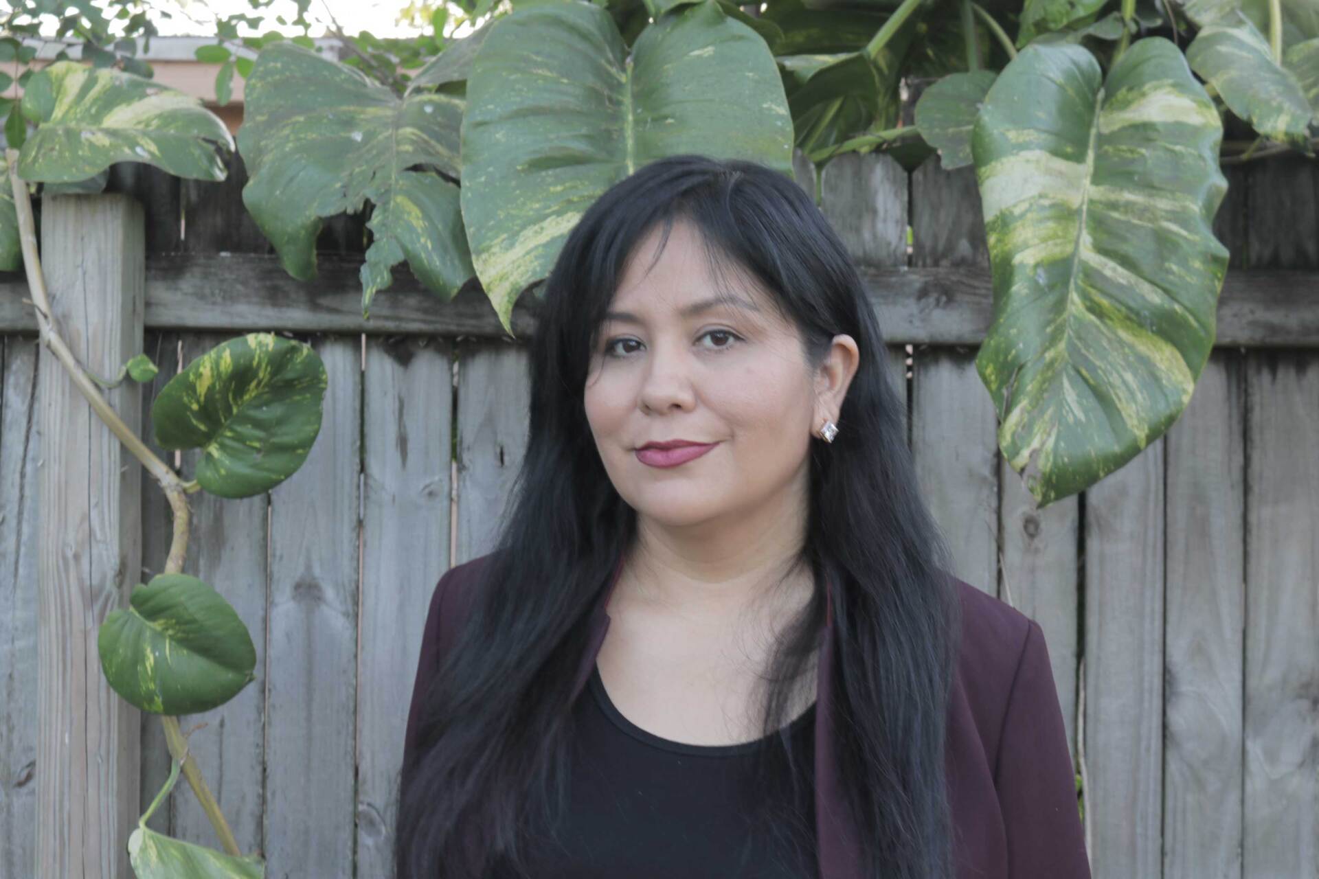 A woman with long black hair is standing in front of a wooden fence.