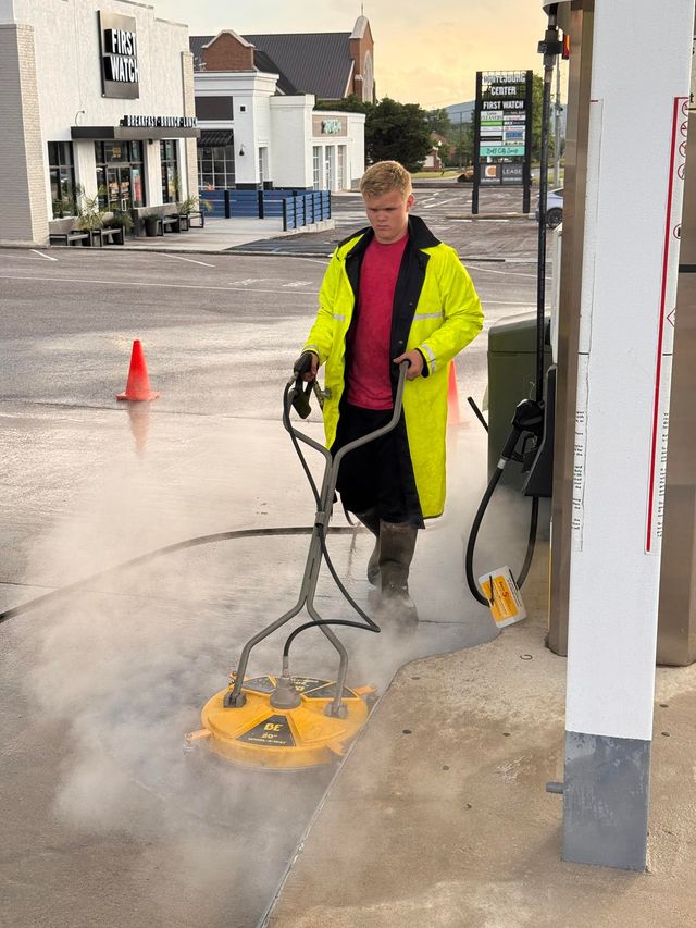 Person in yellow rain gear power washing a concrete surface with a circular cleaner.