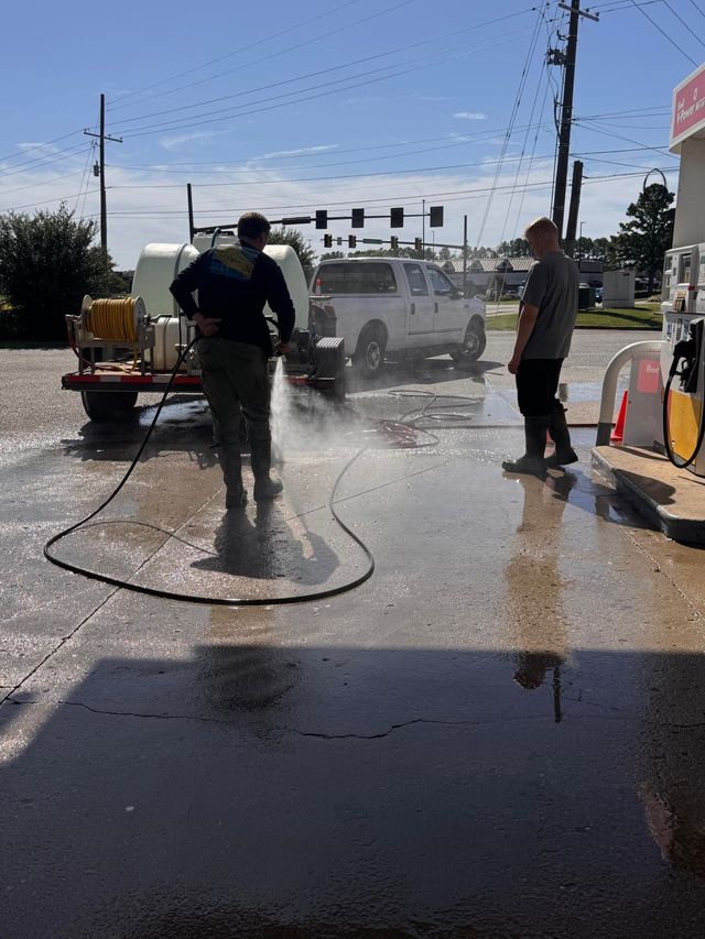 Two people washing a gas station's pavement with water spray. A truck and trailer are parked nearby.