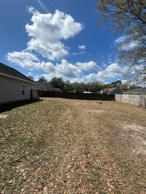 A fenced backyard with dry grass next to a house under a bright blue, partly cloudy sky.