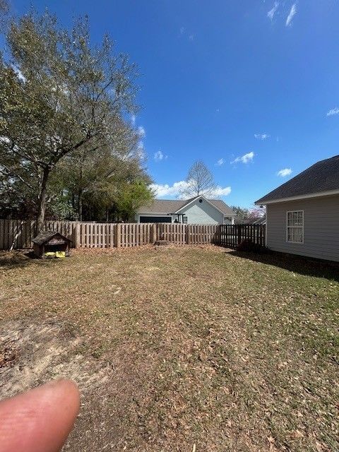 A backyard with dry grass, a wooden fence, a dog house, and a house in the background under a clear blue sky.