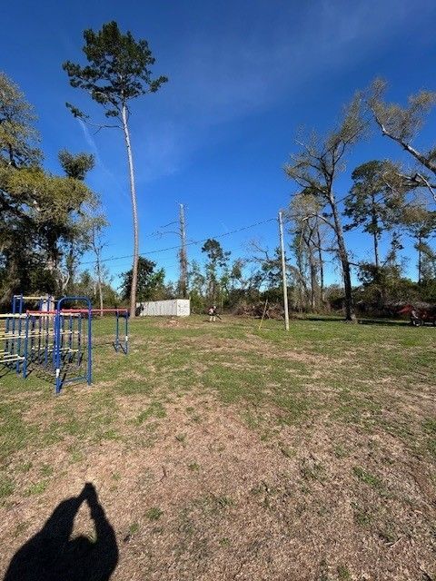A grassy lot with playground equipment on the left, scattered trees, utility poles, and a small shed under a blue sky.