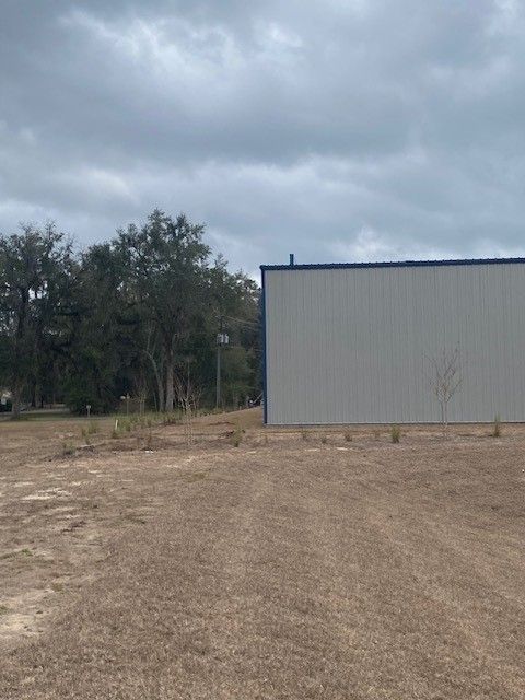 A large, light-colored metal building sits on a dirt lot next to a line of trees under a cloudy sky.