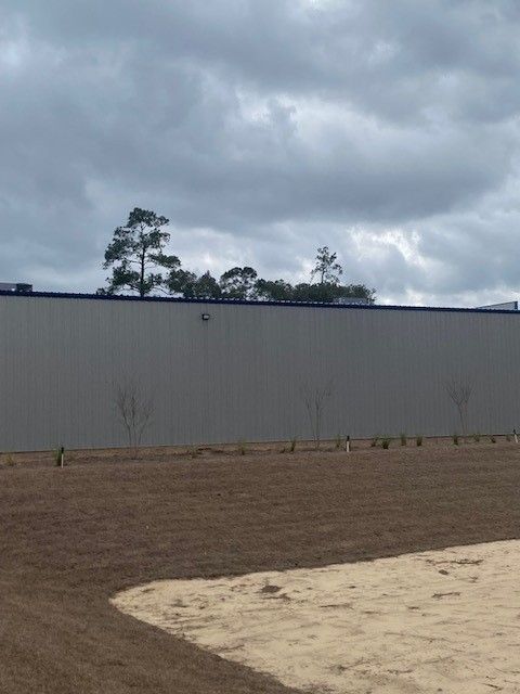 A large, gray industrial building wall stands behind a plot of dirt and sand under a cloudy, overcast sky.