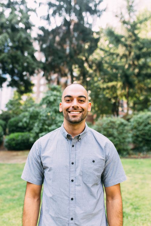 A man in a grey shirt is standing in a park and smiling.