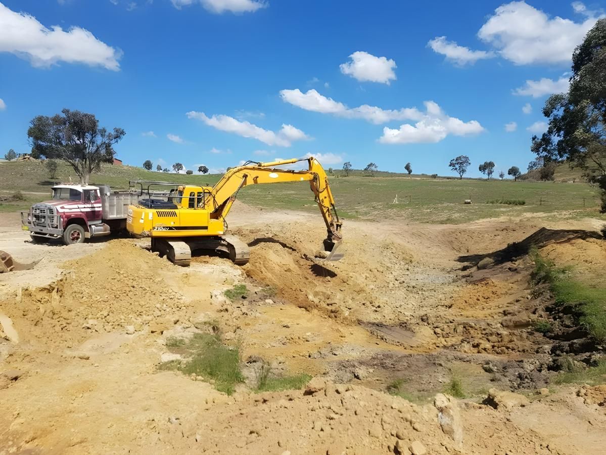 A Yellow Excavator is Digging a Hole in the Dirt Next to a Truck — Hothams In Bathurst, NSW