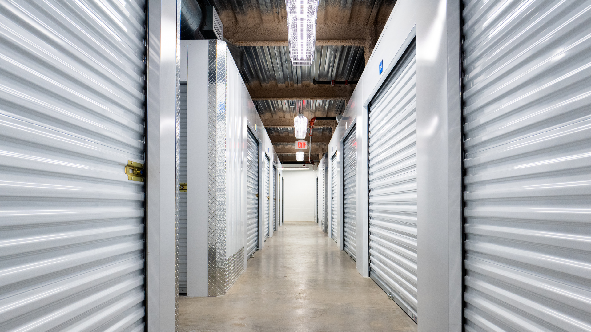 Interior of a storage facility hallway, metal units on both sides, concrete floor, ceiling lights.