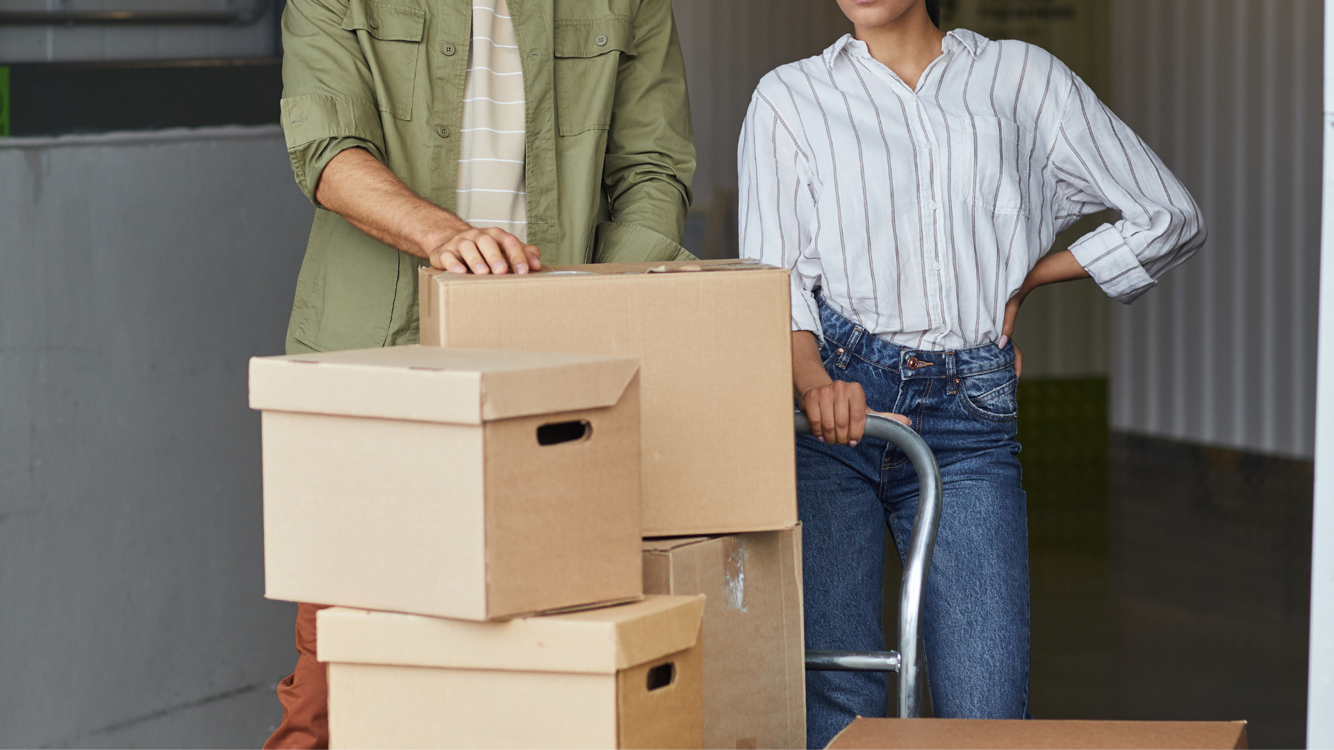 Couple moving boxes in storage unit. Man in green jacket, woman in jeans holding cart.