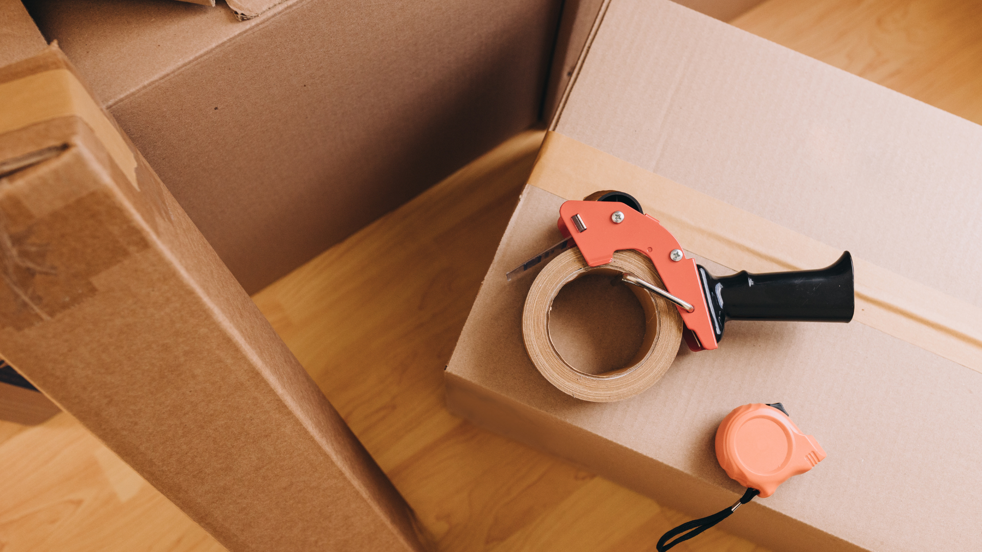 Cardboard boxes being sealed with packing tape, tape dispenser, and measuring tape on wooden floor.