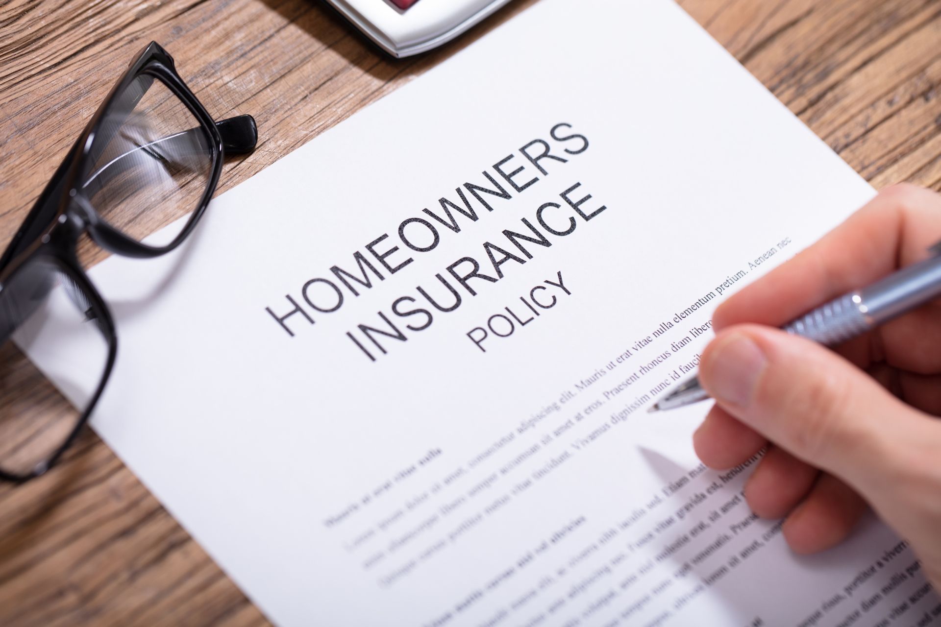 Overhead view of a person's hand filling a homeowner's insurance policy form over a wooden desk.