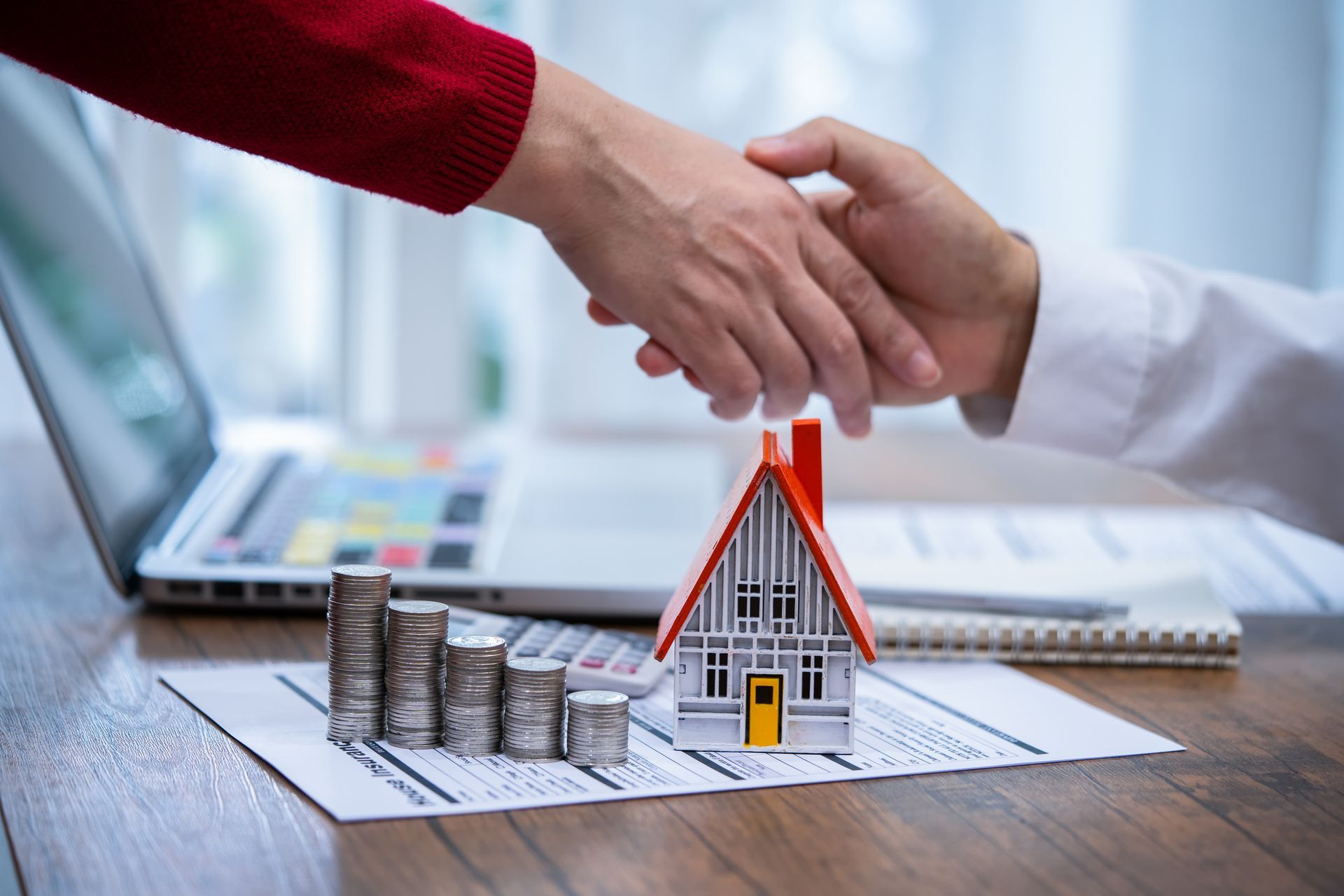 Handshake over house model and stacked coins with laptop in the background.