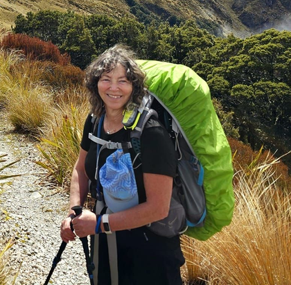 Hiker with green backpack smiling on a mountain trail with tall grasses and rocky hills.