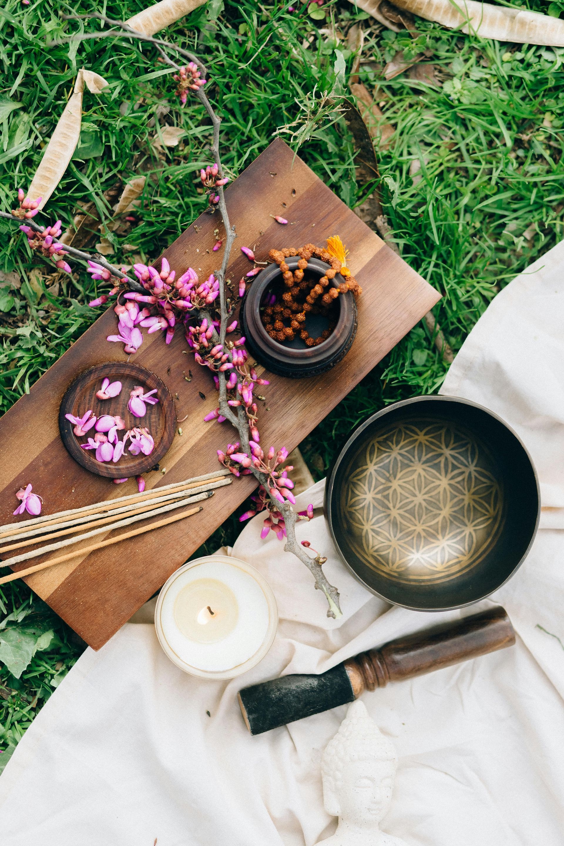 Wooden board with meditation items: singing bowl, candle, incense, beads, flowers, on cloth and grass.