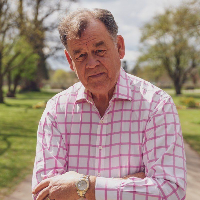 A man wearing a pink and white checkered shirt and a watch is standing in a park.