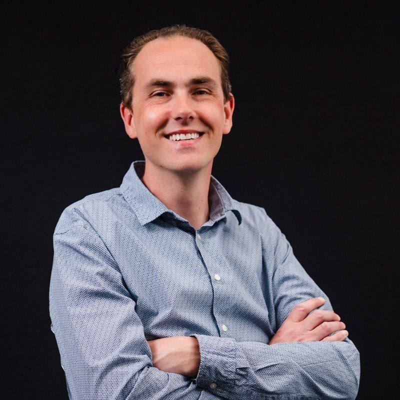 A man in a blue shirt is smiling with his arms crossed.