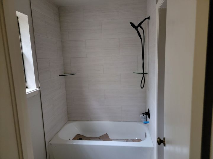 A bathtub with light-colored tile walls, a black shower fixture, and a window to the left in a small bathroom.