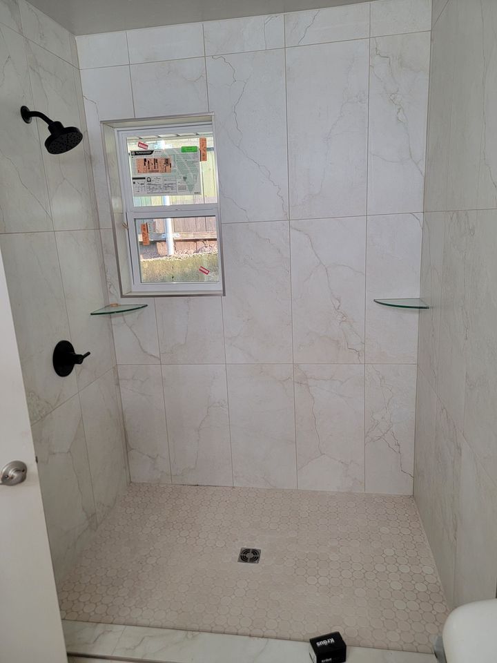 A modern walk-in shower with white marble-patterned tiled walls, a window, black fixtures, and a textured pebble floor.