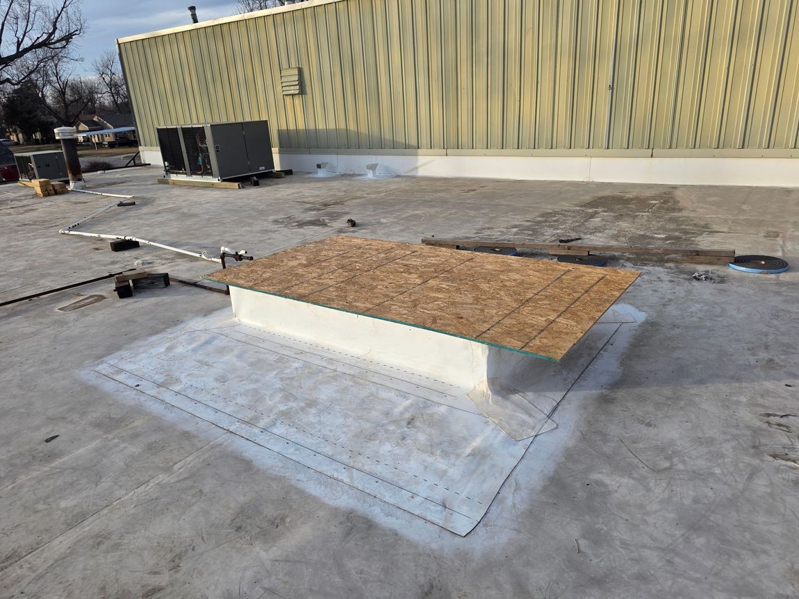 A plywood-covered roof curb sits on a flat commercial roof, surrounded by a white, sealed membrane patch.