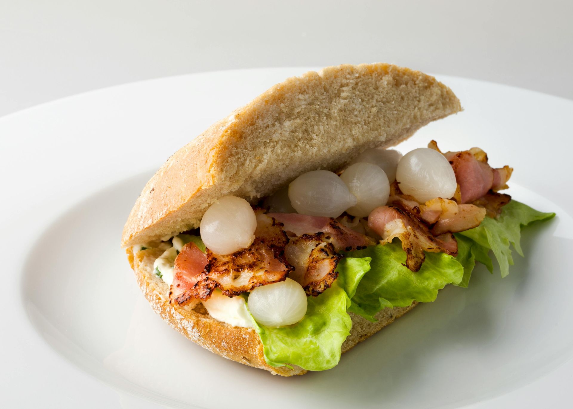A sandwich with bacon, lettuce, and small pickled onions on a rustic bun, served on a white plate.