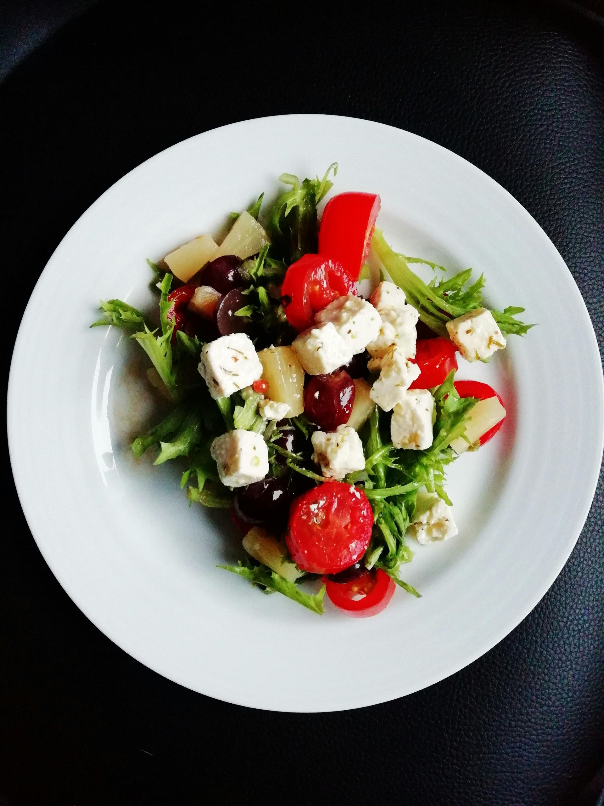 A salad on a white plate containing cubes of white cheese, cherry tomatoes, dark grapes, and green leafy vegetables.