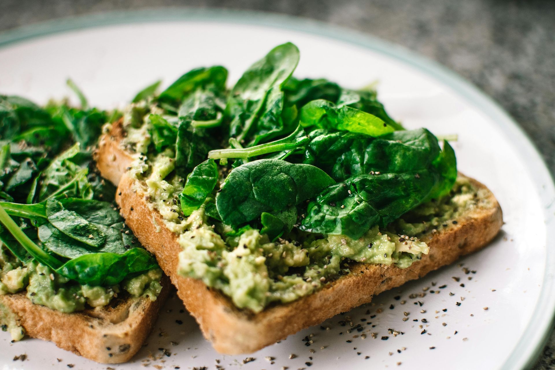 Two slices of toasted bread topped with mashed avocado and fresh spinach leaves, served on a white plate with black pepper.