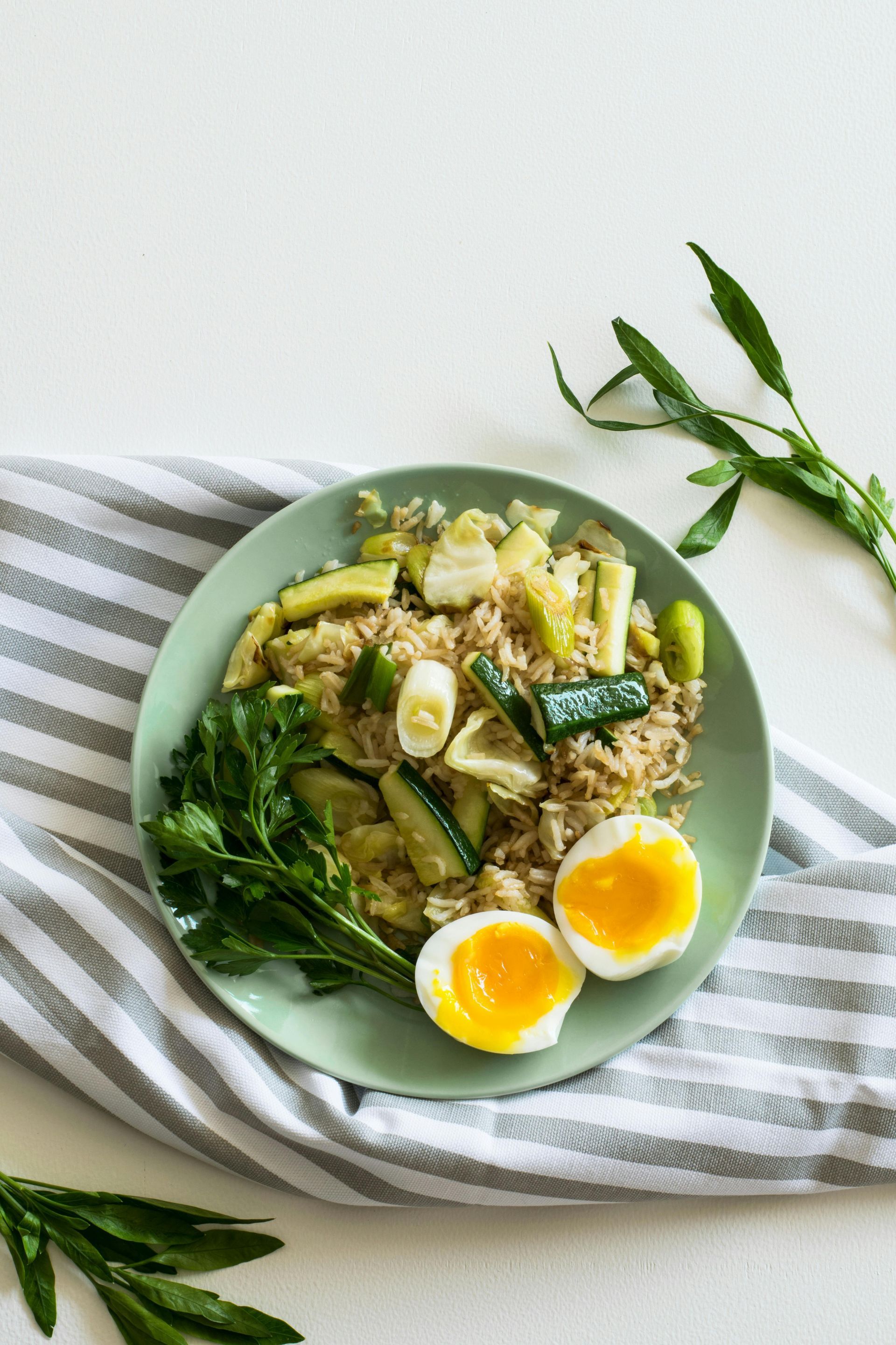 A light green plate of fried rice topped with sliced cucumber and half a soft-boiled egg, set on a striped napkin.