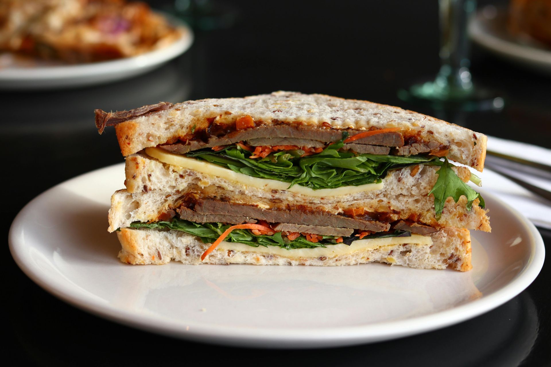A triple-decker sandwich on grain bread, layered with meat, cheese, and fresh greens, centered on a white plate.