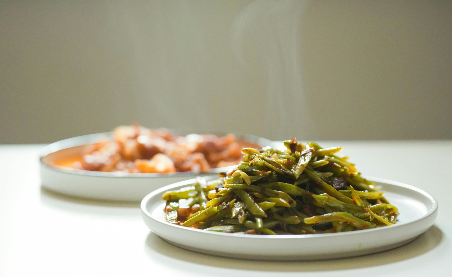 Two plates of hot, steamed food sit on a white surface, featuring stir-fried green beans in the foreground.