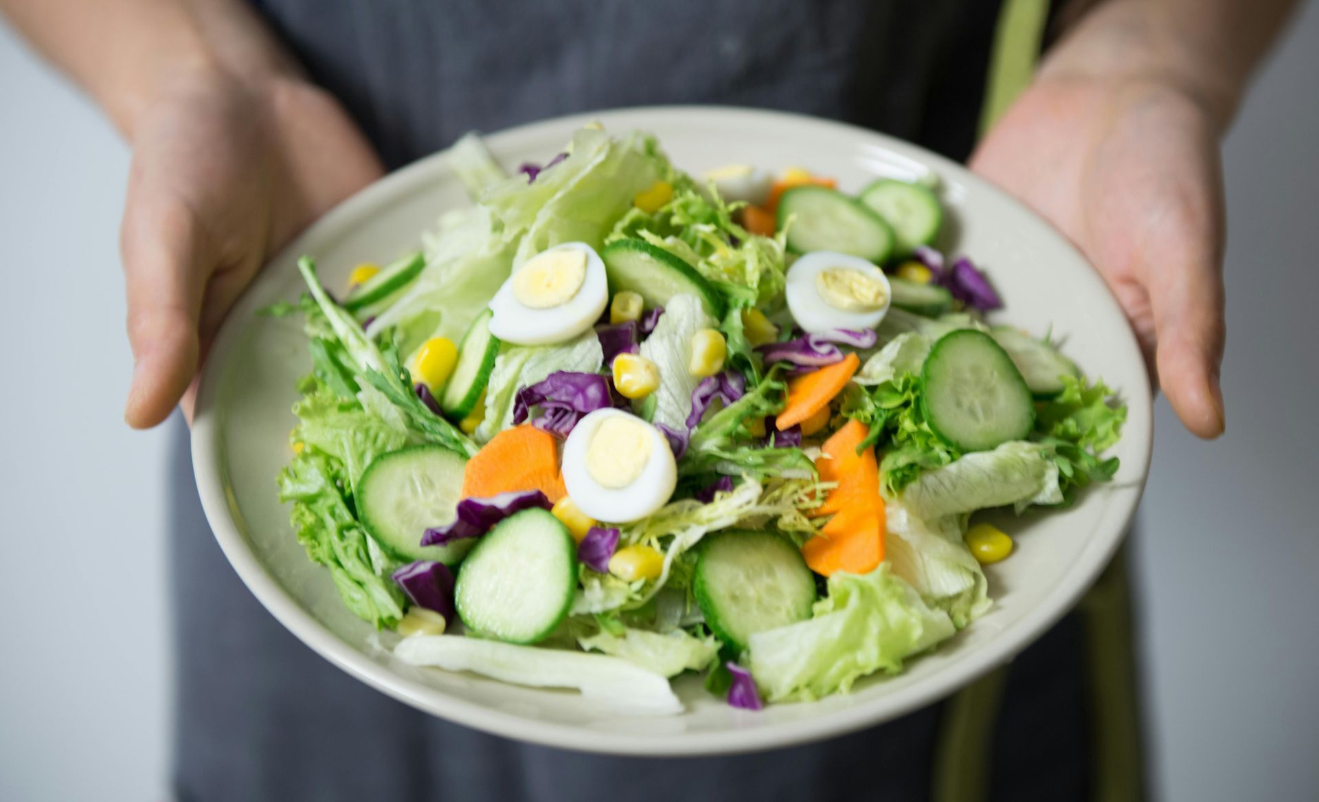 Person holding a plate of fresh salad with lettuce, sliced cucumbers, boiled egg halves, corn, and purple cabbage.