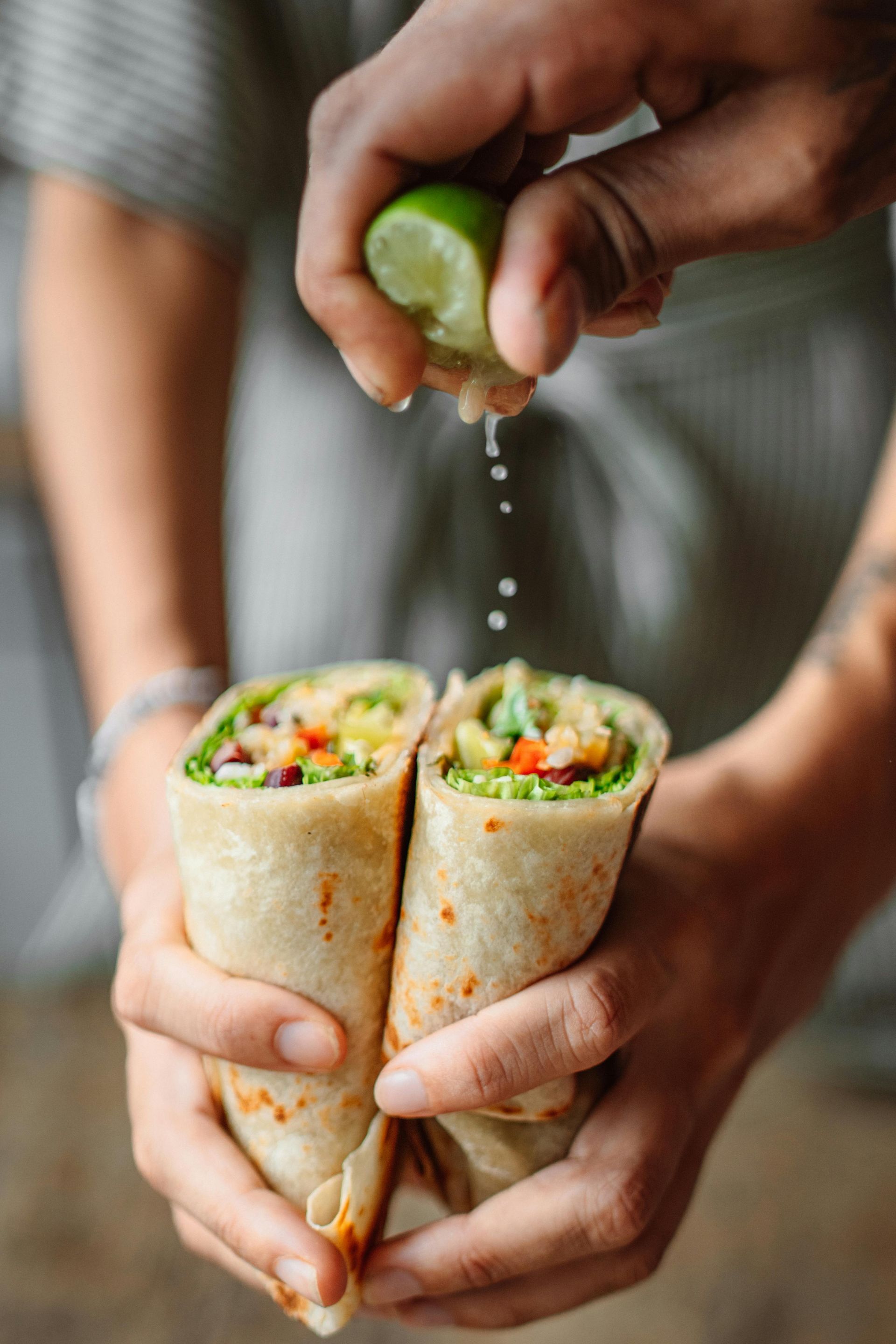 Hands holding two halves of a fresh veggie wrap while squeezing a lime wedge over them, with juice droplets falling.