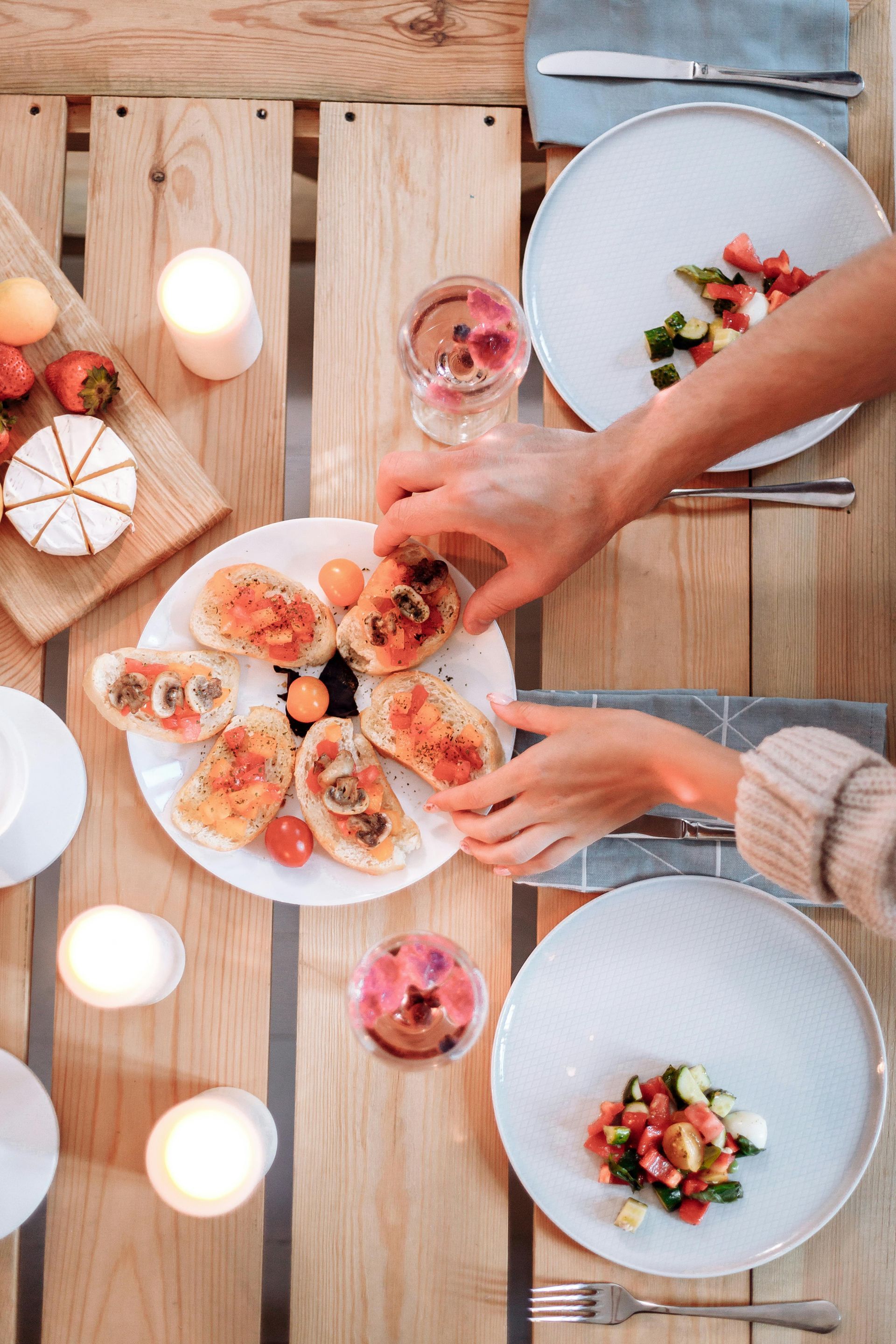 Hands arranging a plate of bruschetta on a wooden table set for dinner with salads, candles, and cheese.