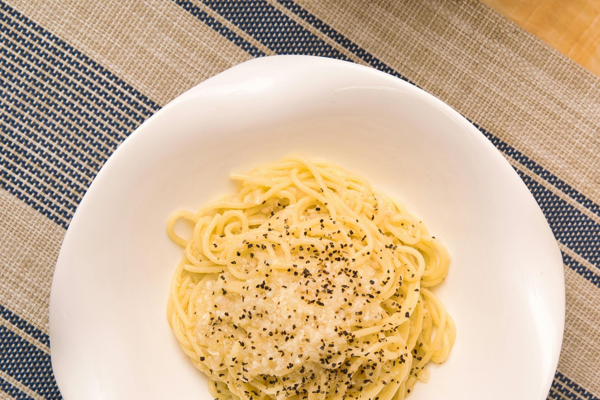 A bowl of pasta topped with cheese and cracked black pepper, set on a blue and white striped cloth surface.