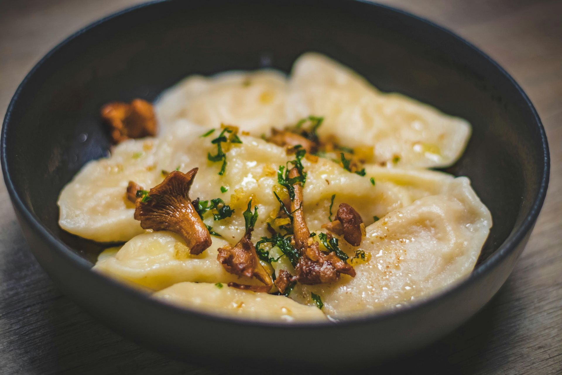 A serving of pierogi topped with sautéed chanterelle mushrooms and fresh herbs in a dark bowl on a wooden surface.