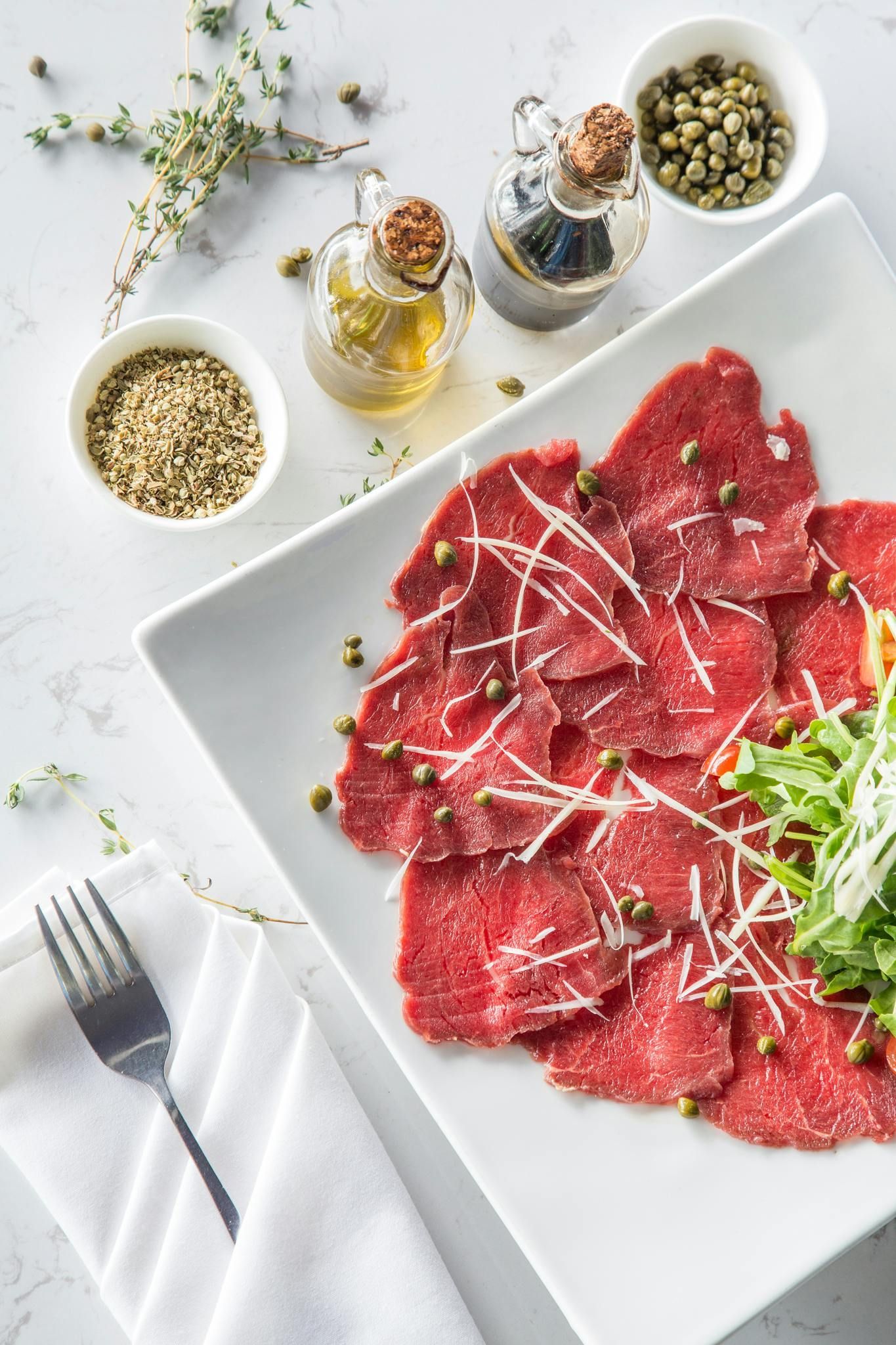 A plate of beef carpaccio topped with arugula, shaved parmesan, and capers, served with olive oil and spices on a table.