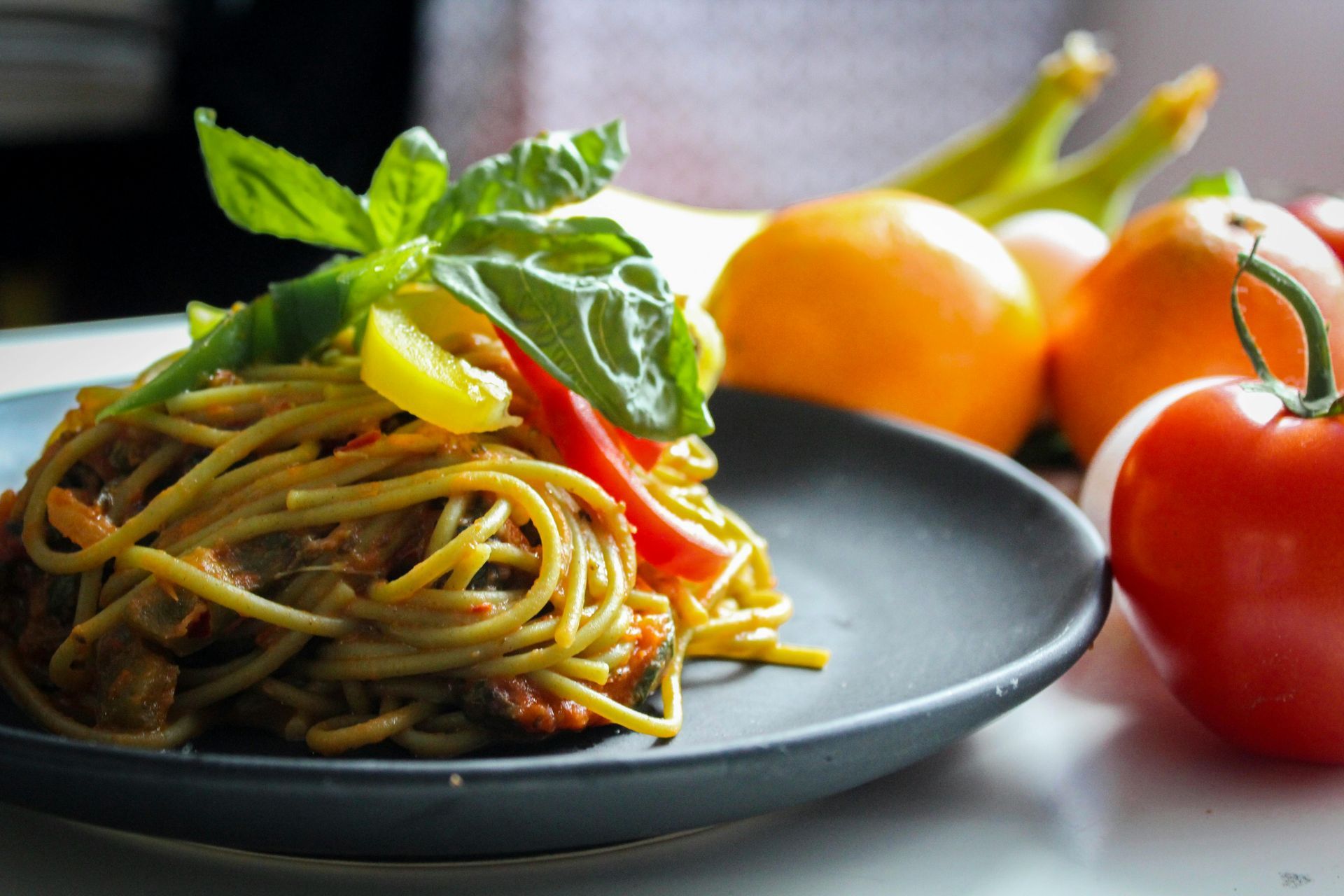 A serving of spaghetti with peppers and basil on a black plate, with fresh tomatoes and oranges in the background.