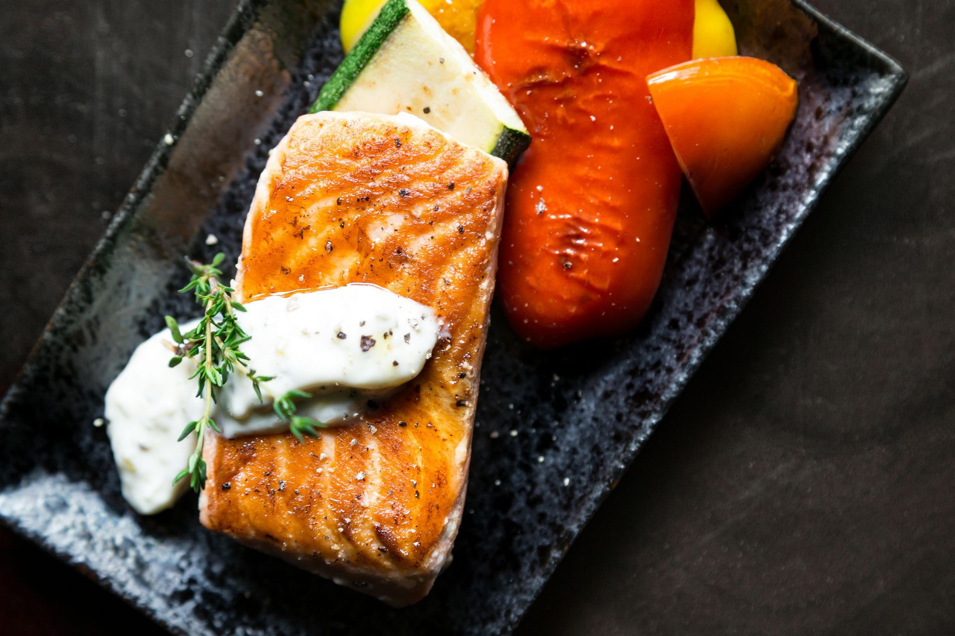 A seared salmon fillet topped with a white cream sauce and herbs, served with roasted vegetables on a dark textured plate.