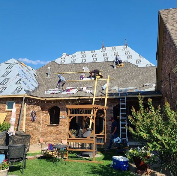 Workers installing shingles on a house roof under a bright blue sky, with a ladder and scaffolding below