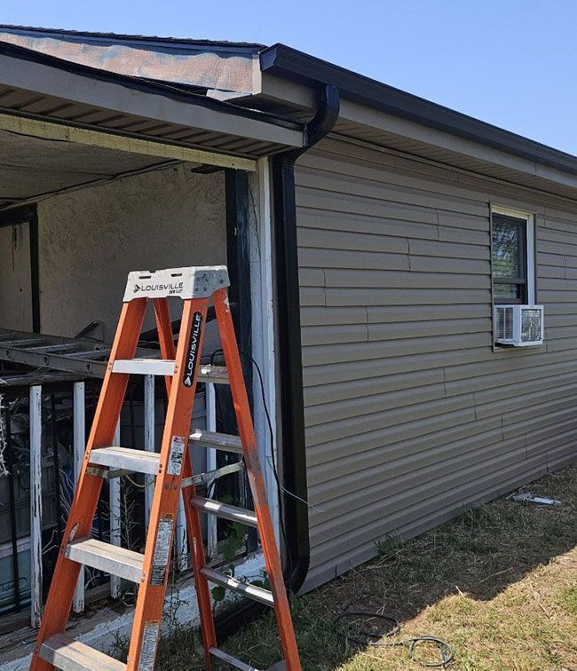 Orange ladder beside a damaged garage opening under a house eave, with beige siding and a window AC unit