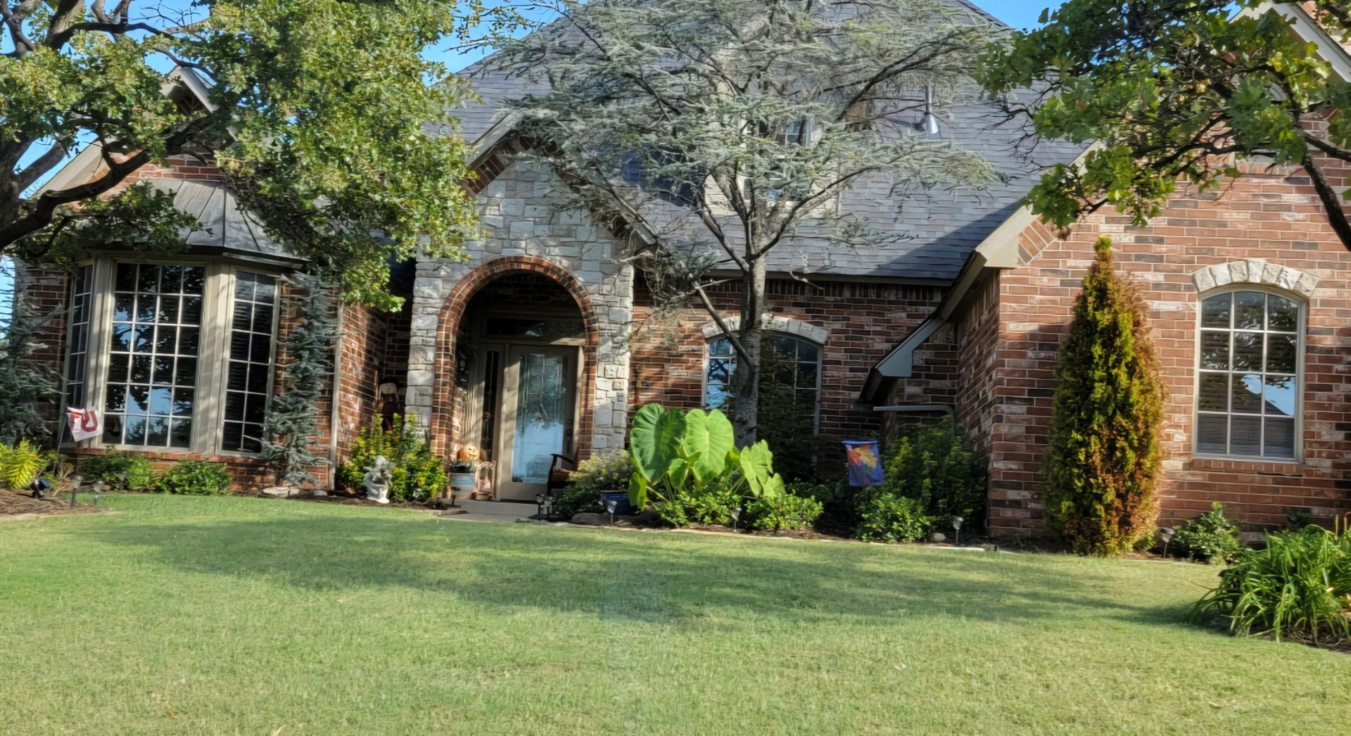 Brick house with arched front entry, large windows, and a green lawn shaded by trees