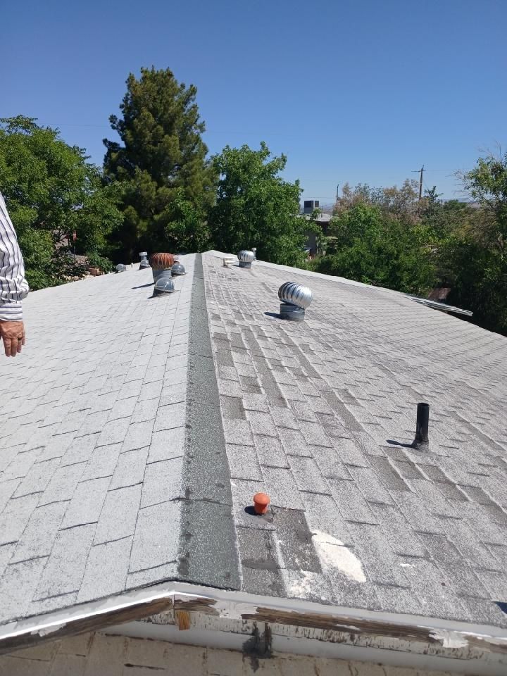 Roof under repair with gray shingles, vents, and one worker visible against trees and blue sky